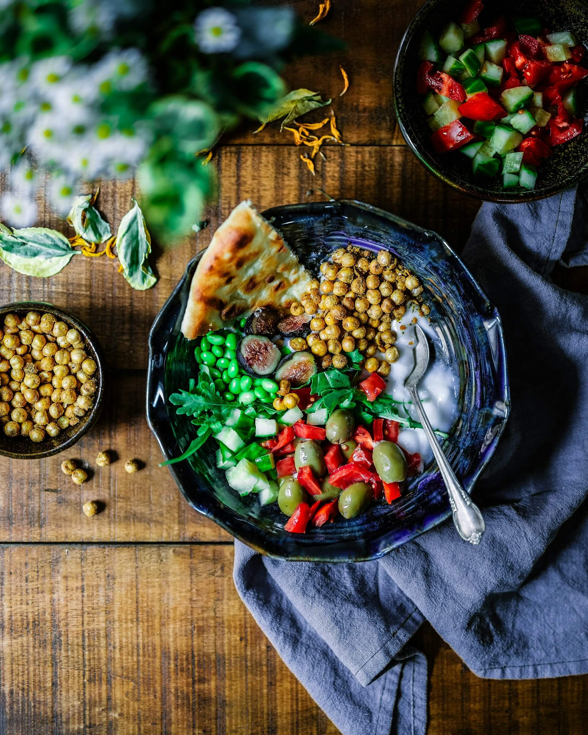 A bowl filled with delicious looking food taken from above. In the background of the picture you can see a rough wooden table with two smaller bowls and a blue kitchen towel.