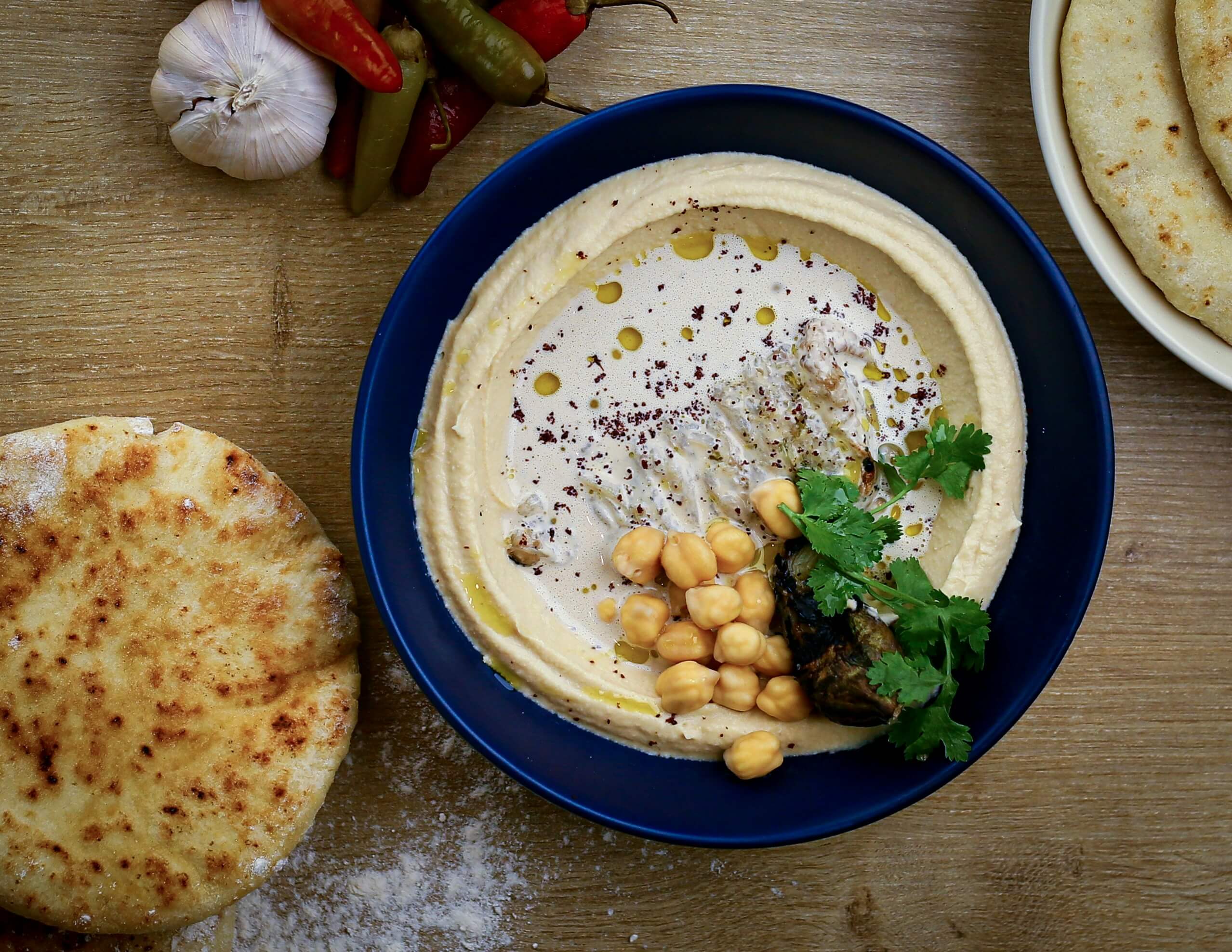 A bright blue bowl filled with hummus taken from above. On top of the hummus, there are a few chickpeas and a sprig of coriander. Next to the bowl there are pita bread, garlic and some chilli.