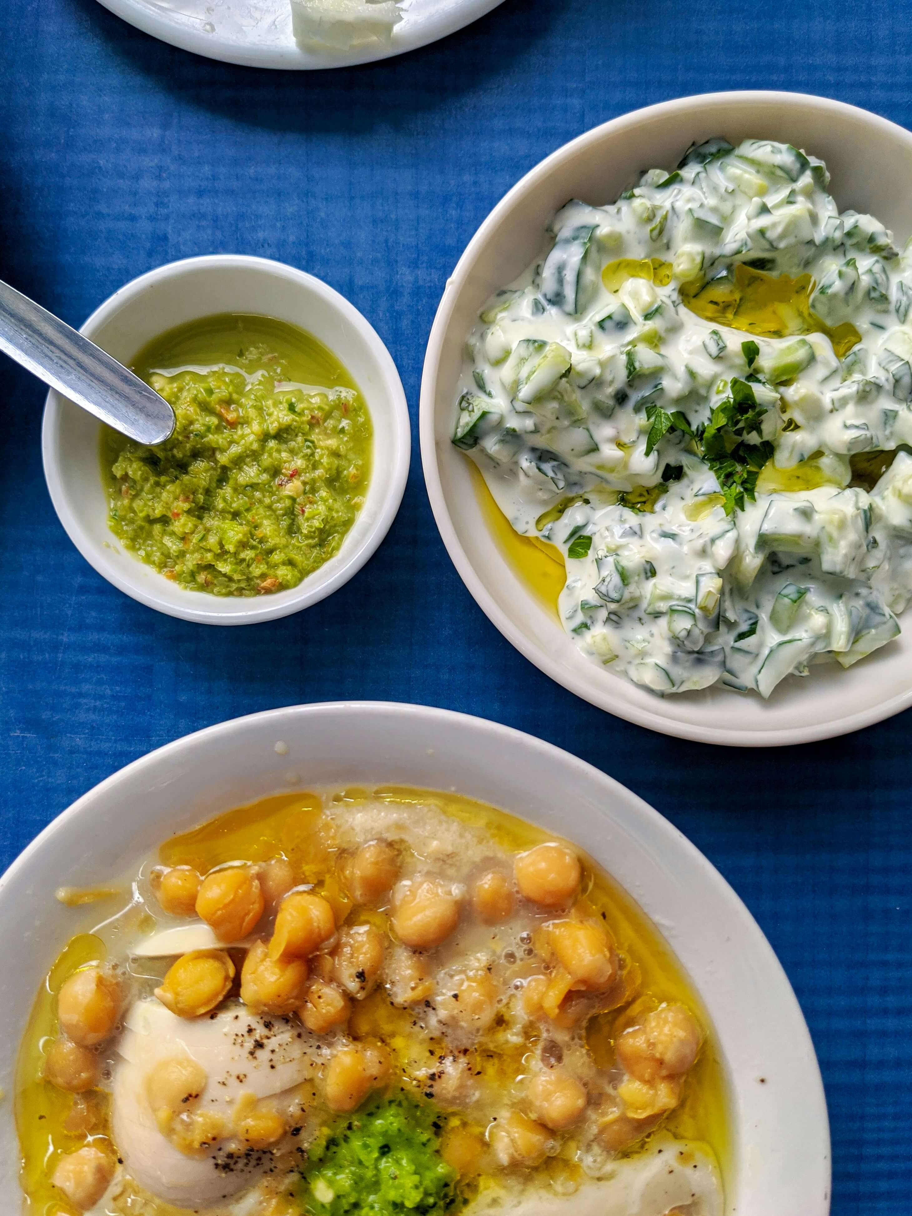 Taken from above we can see three bowls, varying in size filled with food. The smallest one is filled with a green sauce, next to that one sits a bowl filled with tzatziki and beneath the two bowls sits the lagrgest one filled with a chickpea stew. Underneath the tree bowls, is a bright blue tablecloth.